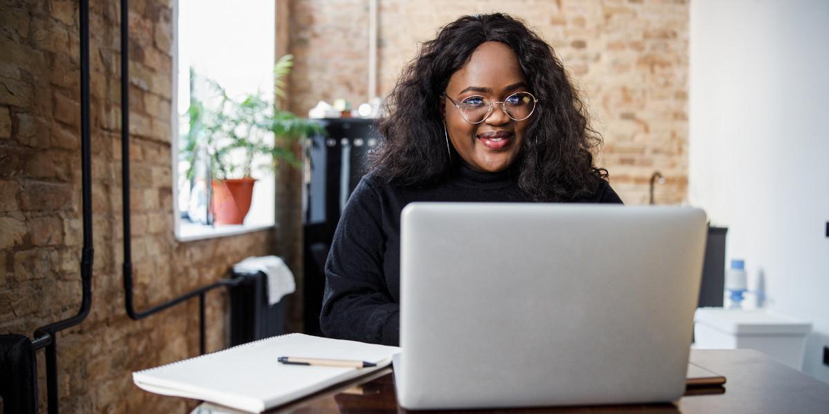 A Black woman with glasses sits at a desk with an open laptop and a notepad.